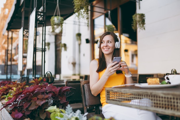 Bella donna in una caffetteria all'aperto di strada seduta al tavolo, ascolta musica in cuffia, usa il telefono cellulare, rilassati al ristorante nel tempo libero