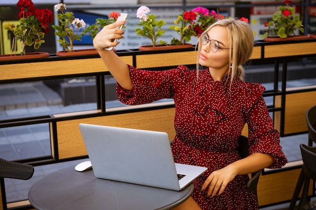 Bella donna in vestito rosso e bicchieri seduti nella caffetteria sulla strada con il computer portatile fa selfie sul telefono