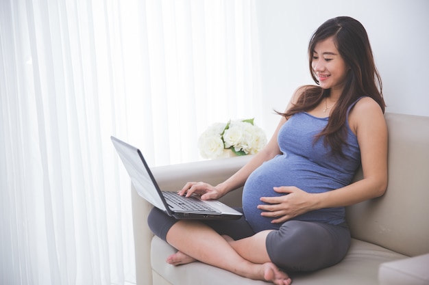 Bella donna incinta che controlla un pc del computer portatile mentre sedendosi sul sofà