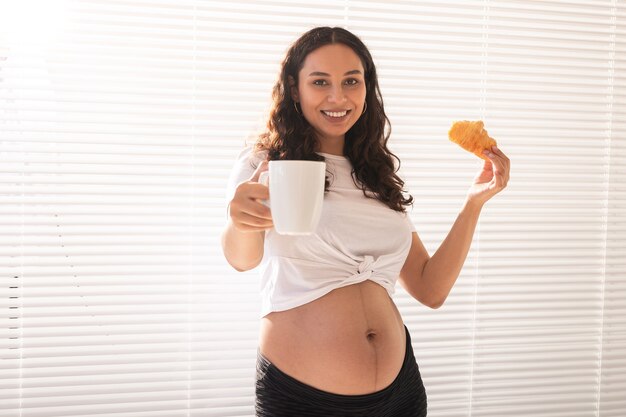 Bella donna incinta che tiene croissant e tazza di caffè nelle sue mani durante la colazione del mattino