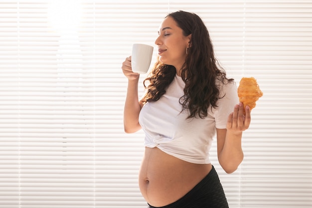Bella donna incinta che tiene croissant e tazza di caffè nelle sue mani durante la colazione del mattino