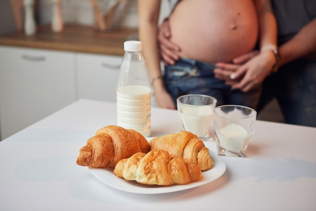 Bella donna incinta e suo marito che godono di mangiare croissant al cioccolato con latte