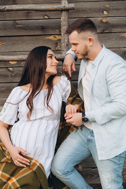 Bella donna incinta e suo marito in posa vicino a un edificio in legno. Le coppie felici alla moda si amano. Moda e stile rustico.