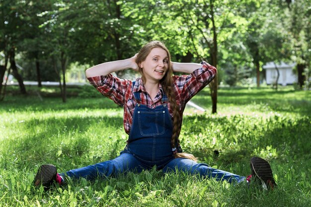 Bella donna incinta in tuta di jeans seduta sull'erba nel parco
