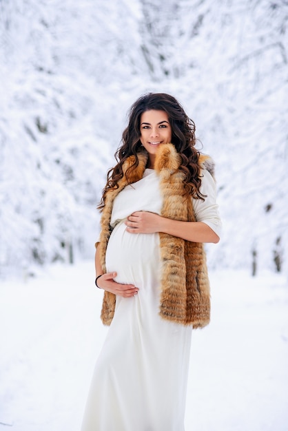 Bella donna incinta in un lungo abito leggero e cappotto tiene delicatamente il suo stomaco con le mani in un