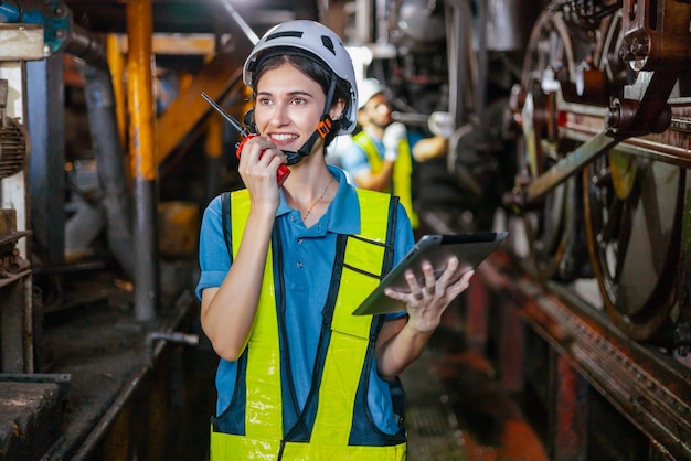 Bella donna ingegnere in fabbrica che parla alla radio indossando casco bianco e abiti da lavoro e mano che tiene tablet sul posto di lavoro