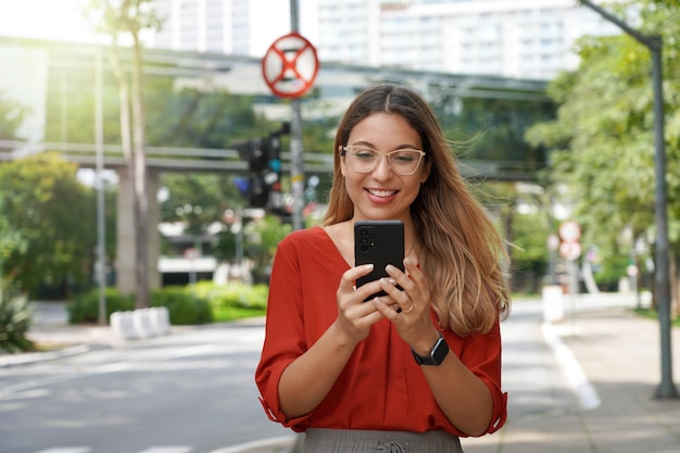 Bella donna intelligente con gli occhiali che utilizza il telefono cellulare che cammina nella moderna metropoli sostenibile di San Paolo in Brasile