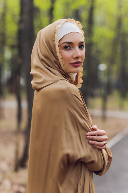 Bella donna islamica in un vestito musulmano che sta sugli alberi di autunno della foresta del fondo della strada del parco di estate