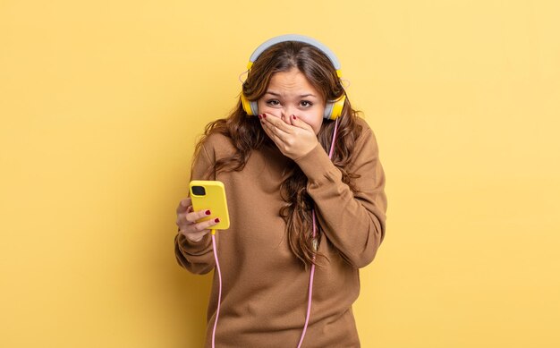 Bella donna ispanica che copre la bocca con le mani con uno shock. concetto di cuffie e smartphone