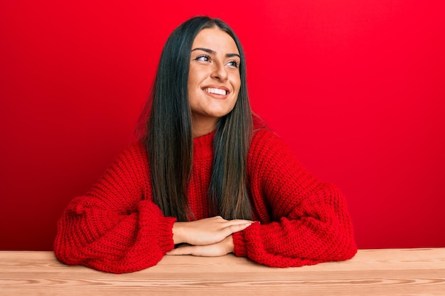 Bella donna ispanica che indossa abiti casual seduta sul tavolo che distoglie lo sguardo a lato con il sorriso sul viso, espressione naturale. ridendo fiducioso.