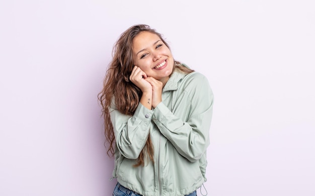 Bella donna ispanica che si sente innamorata e sembra carina, adorabile e felice, sorride romanticamente con le mani accanto al viso