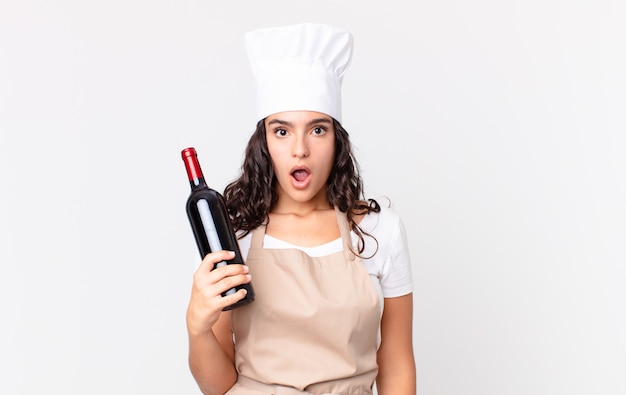 Bella donna ispanica chef che sembra molto scioccata o sorpresa e tiene in mano una bottiglia di vino
