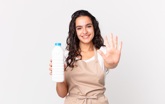 Bella donna ispanica chef sorridente e dall'aspetto amichevole, che mostra il numero cinque e tiene in mano una bottiglia di latte