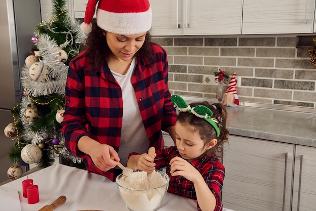 Bella donna ispanica, mamma amorevole felice e la sua amata figlia carina in abito a scacchi rosso che si divertono insieme, preparando l'impasto per cucinare i panifici di Natale a casa cucina. 25 dicembre