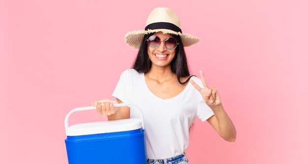 Bella donna ispanica sorridente e dall'aspetto amichevole, che mostra il numero due con un frigorifero portatile da picnic