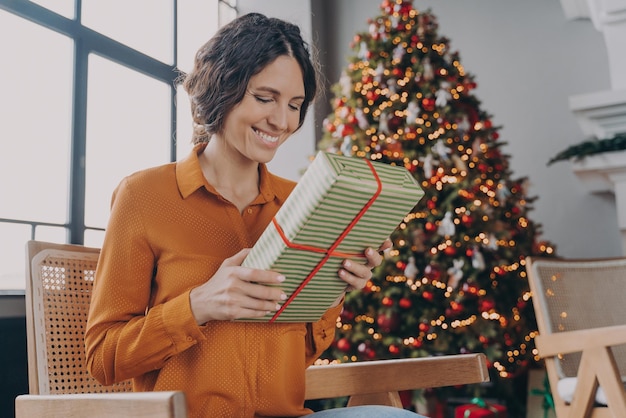 Bella donna italiana felice seduta con un regalo accanto all'albero di Natale decorato a festa a casa