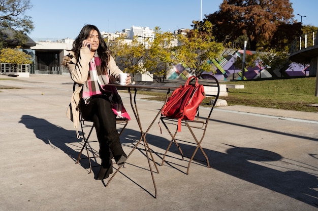 Bella donna latina che parla al cellulare mentre beve un caffè sulla terrazza di un bar in una giornata di sole. Relax e tempo libero, comunicazione, concetto di tecnologia.