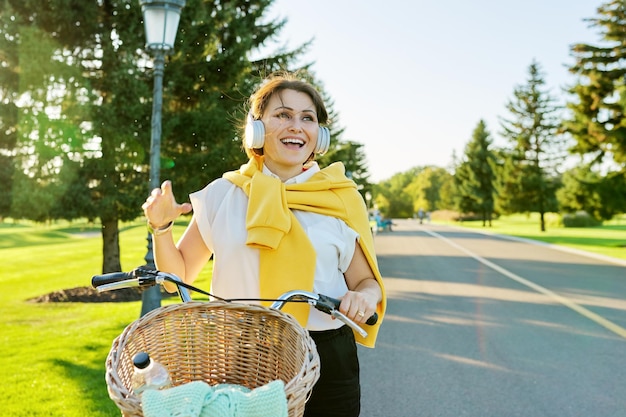 Bella donna matura attiva con la bici sulla strada nel parco Donna sorridente felice di mezza età con le cuffie che ascolta lo spazio della copia della musica