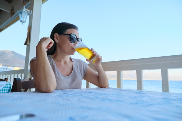 Bella donna matura che riposa seduto in caffè sul mare a bere un bicchiere di birra fredda