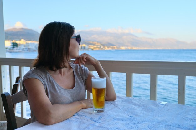 Bella donna matura che riposa seduto in caffè sul mare con un bicchiere di birra fredda