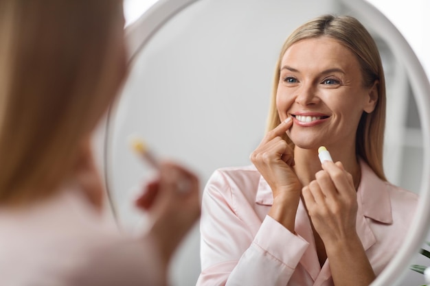 Bella donna matura che si guarda allo specchio e si applica balsamo per le labbra idratante