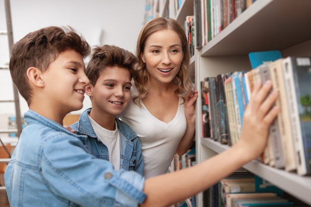 Bella donna matura e figli gemelli che scelgono un libro dallo scaffale della biblioteca