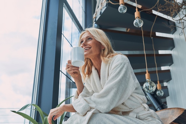 Bella donna matura in accappatoi che beve caffè e sorride mentre è seduta vicino alla finestra a casa