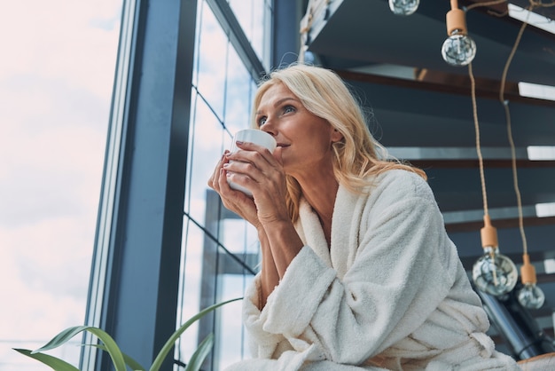 Bella donna matura in accappatoi che beve caffè e sorride mentre si trova vicino alla finestra a casa