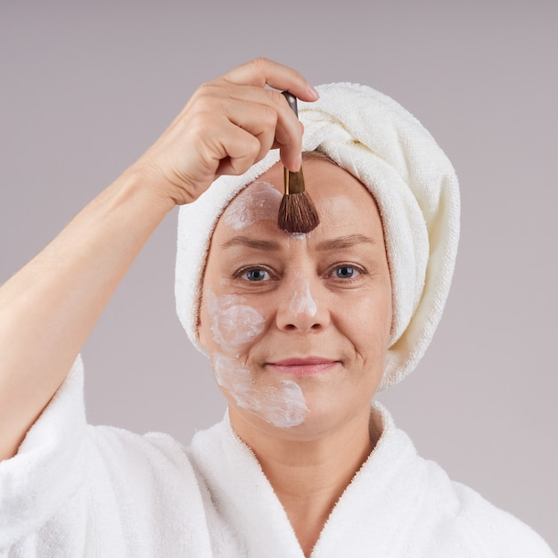 Bella donna matura in camice bianco che applica una maschera nutriente al viso. Isolare su un muro grigio.