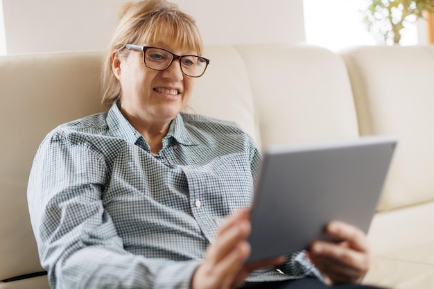 Bella donna matura in occhiali sta usando una tavoletta digitale e sorride mentre era seduta sul divano di casa