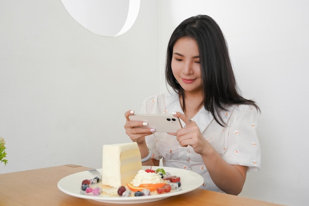 Bella donna millenaria asiatica che scatta una foto di deliziosi dessert con il suo smartphone nella caffetteria