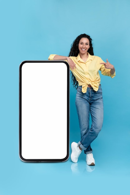 Bella donna millenaria che offre mockup per la nuova app mobile sullo schermo gigante dello smartphone in blu