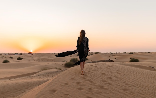 Bella donna misteriosa in abito lungo nero arabo tradizionale si trova nel deserto al tramonto