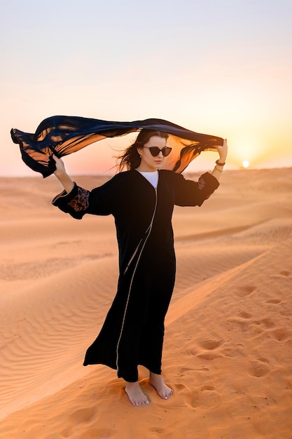 Bella donna misteriosa in abito lungo nero arabo tradizionale si trova nel deserto al tramonto
