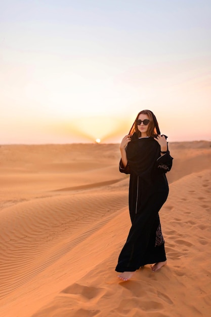 Bella donna misteriosa in abito lungo nero arabo tradizionale si trova nel deserto al tramonto