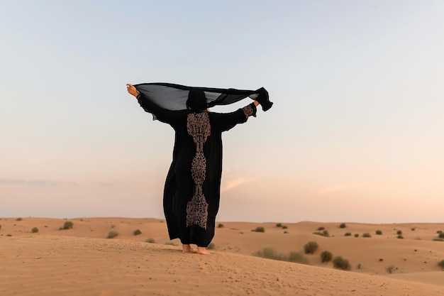 Bella donna misteriosa in abito lungo nero arabo tradizionale si trova nel deserto al tramonto