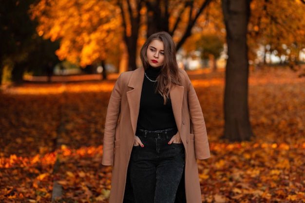 Bella donna modello con labbra rosse in un cappotto beige alla moda e maglione cammina nel parco autunnale con fogliame arancione colorato