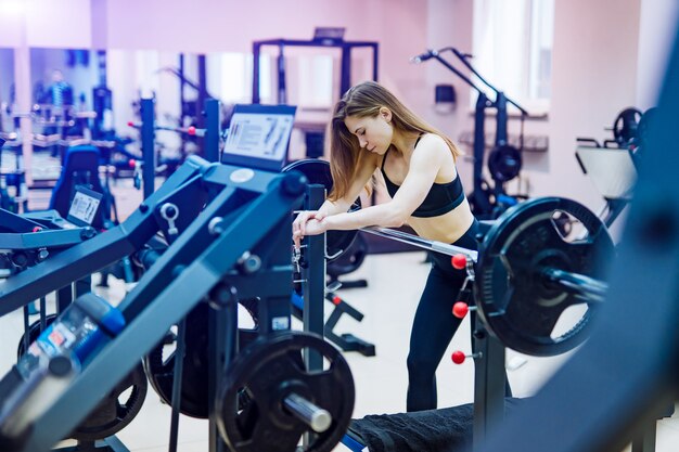 Bella donna muscolare di forma fisica in vestiti sportivi neri che si appoggiano simulatore con il bilanciere alla palestra.