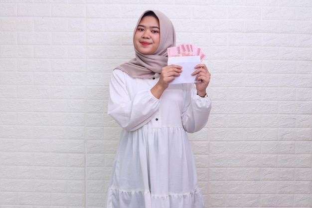 Bella donna musulmana asiatica che indossa l'hijab che mostra dei soldi sulla busta come regalo di eid al fitr