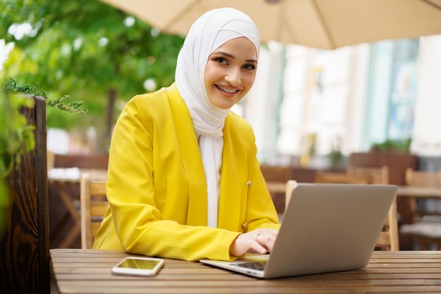 Bella donna musulmana sorridente con il computer portatile al caffè