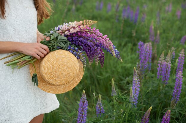 Bella donna nel campo del lupino. Ragazza che tiene un mazzo di lupini e cappello. Estate.