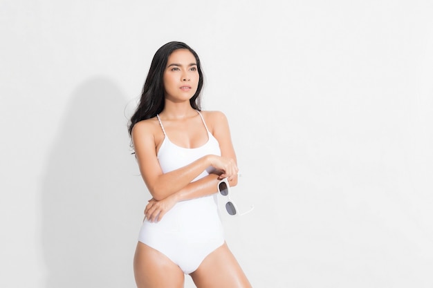 Bella donna nella felicità che indossa il vestito bianco dal costume da bagno e che tiene gli occhiali da sole bianchi post nel tiro dello studio di moda estivo di concetto isolato su priorità bassa bianca.