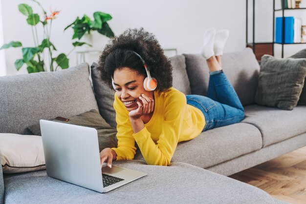 Bella donna nera adulta a casa che ascolta musica online con computer portatile e cuffie - Bella ragazza nera che lavora in remoto online a casa