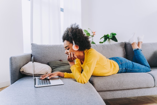 Bella donna nera adulta a casa che ascolta musica online con computer portatile e cuffie - Bella ragazza nera che lavora in remoto online a casa