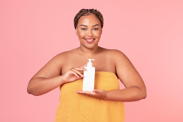 Bella donna obesa nera avvolta in un asciugamano che tiene una bottiglia con una lozione per il corpo idratante che usa un prodotto per la cura della pelle