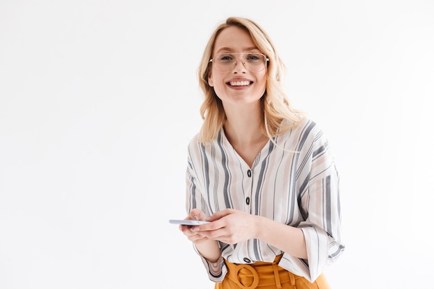 Bella donna ottimista che indossa occhiali sorride alla macchina fotografica e digita sullo smartphone isolato sopra il muro bianco