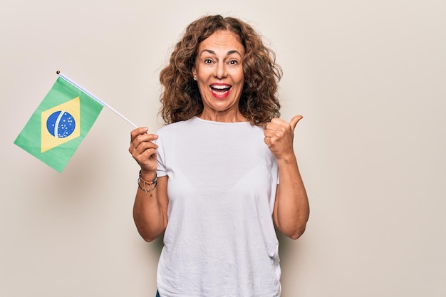 Bella donna patriottica di mezza età che tiene bandiera brasiliana su sfondo bianco isolato che punta il pollice fino al lato sorridente felice con la bocca aperta