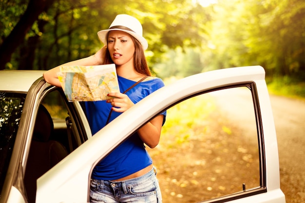 Bella donna persa confusa in piedi appoggiata all'auto nella foresta e preoccupata guardando una mappa.