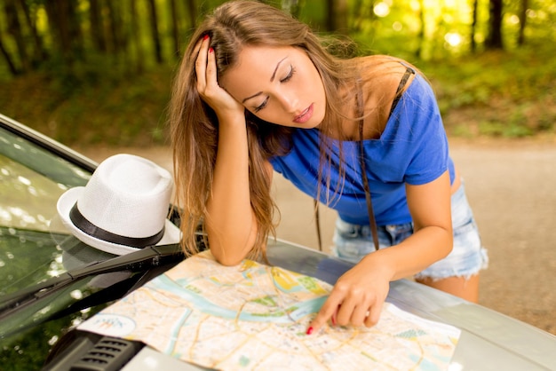 Bella donna persa confusa in piedi appoggiata all'auto nella foresta e preoccupata guardando una mappa.