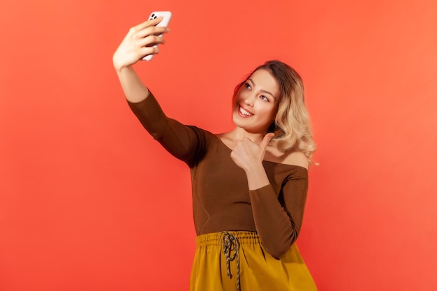 Bella donna positiva che fa selfie o registra video su smartphone per i suoi abbonati che mostrano i pollici in su chiedendo di mettere mi piace ai suoi post Scatto in studio al coperto isolato su sfondo arancione